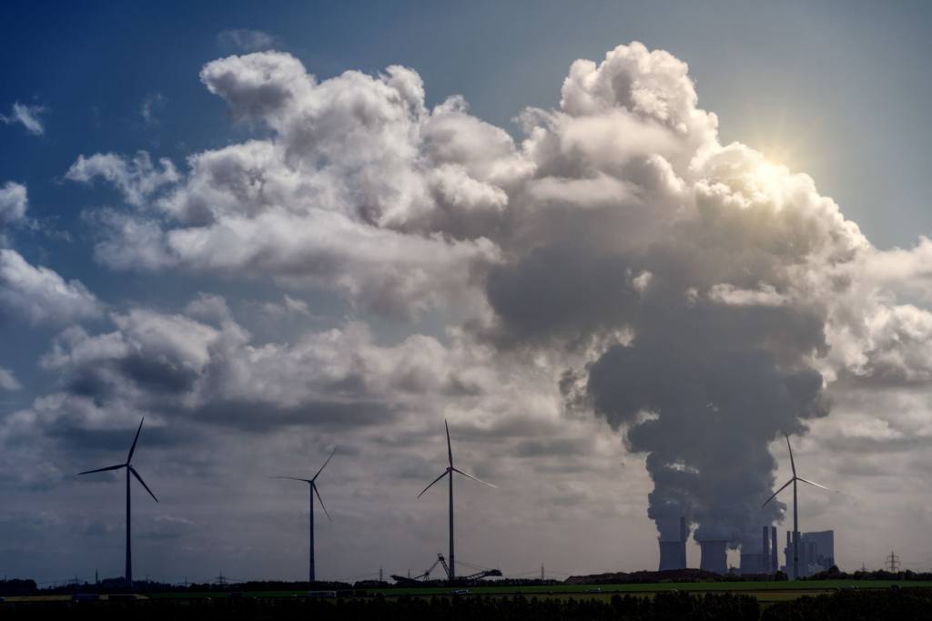 Wind turbines standing beside an industrial cooling tower releasing a large steam plume into a blue sky