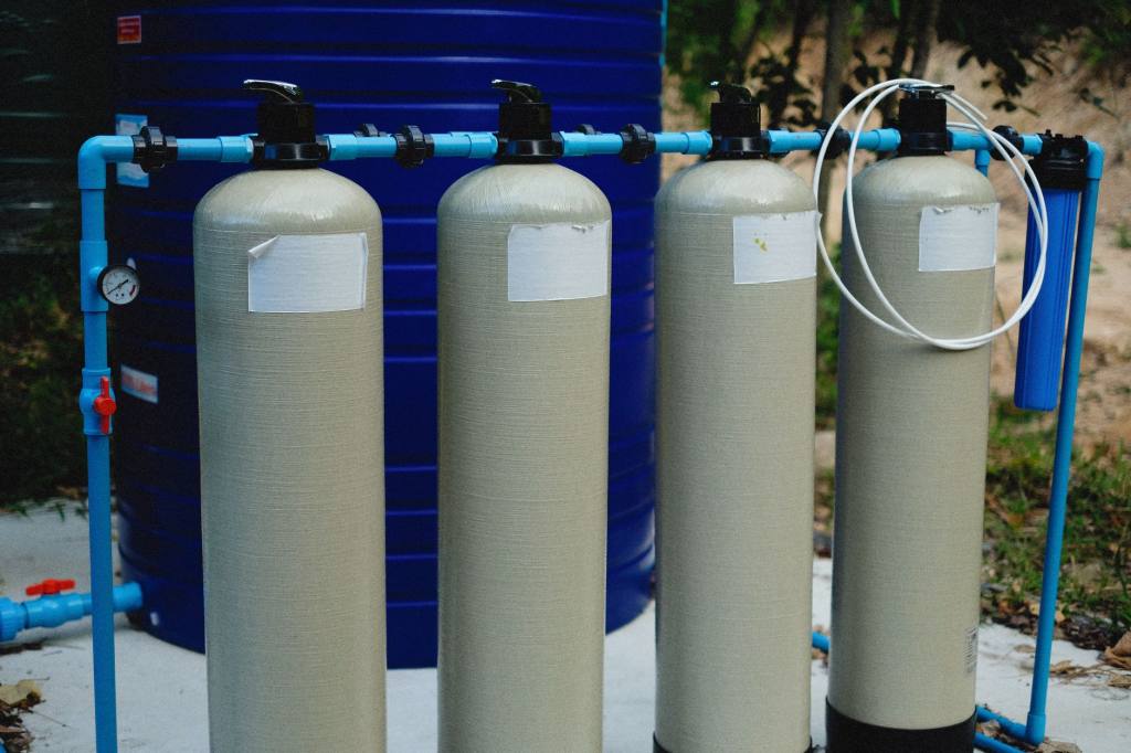 Row of four cylindrical filter vessels connected by blue pipework with a pressure gauge and valves at an outdoor water treatment installation
