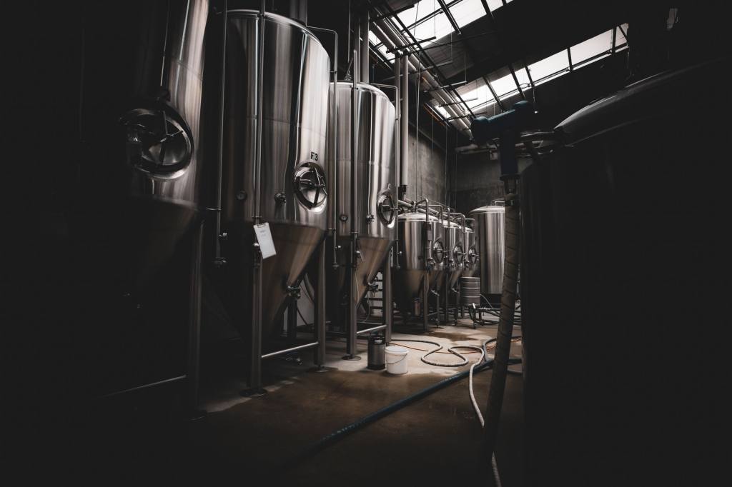 Tall stainless steel conical process tanks with pipework and valves in a dimly lit industrial hall