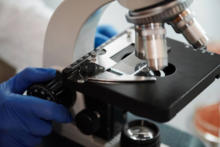 SENZAIT Analytical research — a gloved hand adjusts the graduated stage of a microscope over a sample slide.