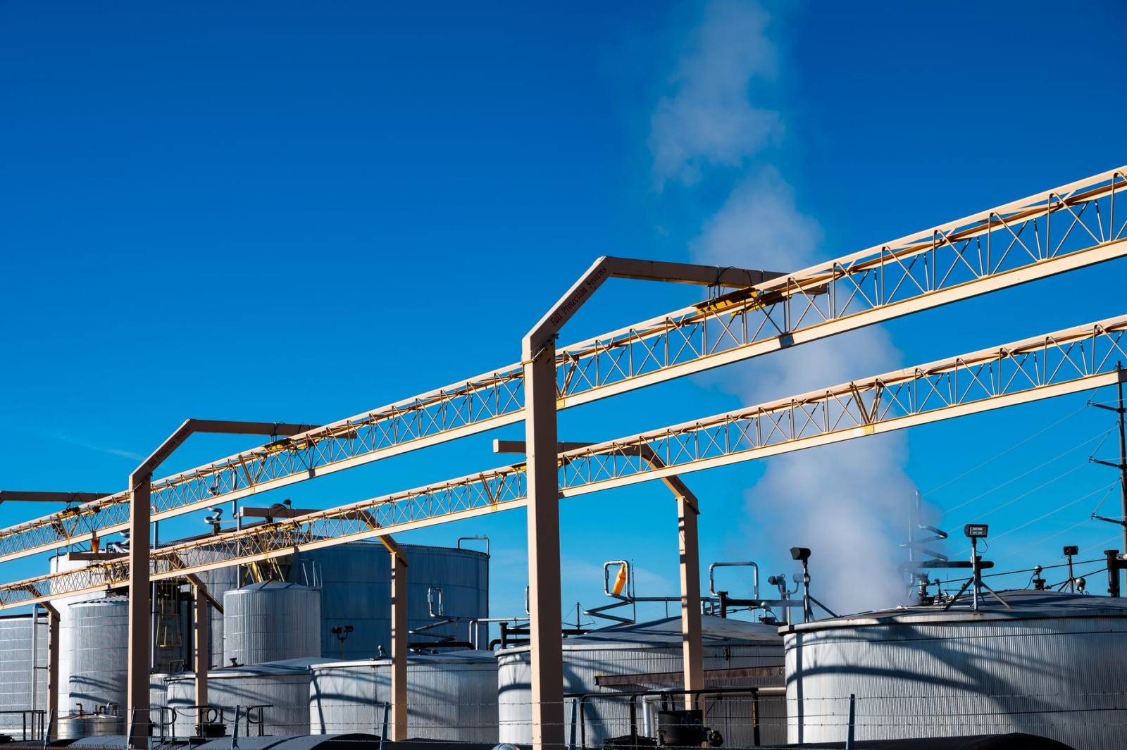 SENZAIT Industries served — overhead pipe racks and storage tanks of a chemical plant against a clear sky, a plume rising behind them.