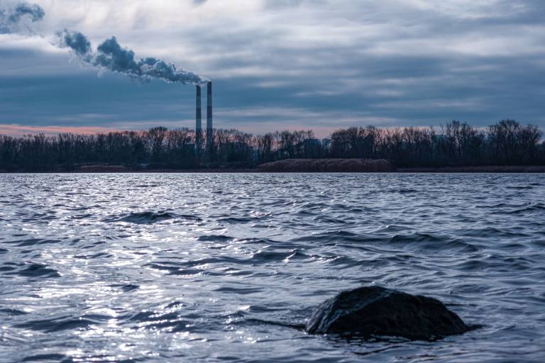 SENZAIT Environment — two industrial stacks and their plume behind a tree line, seen across open river water under a heavy sky.