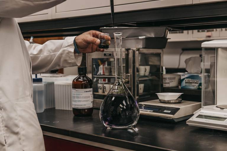 SENZAIT Laboratory validation — a scientist pours a measured sample into a volumetric flask beside an analytical balance at the bench.